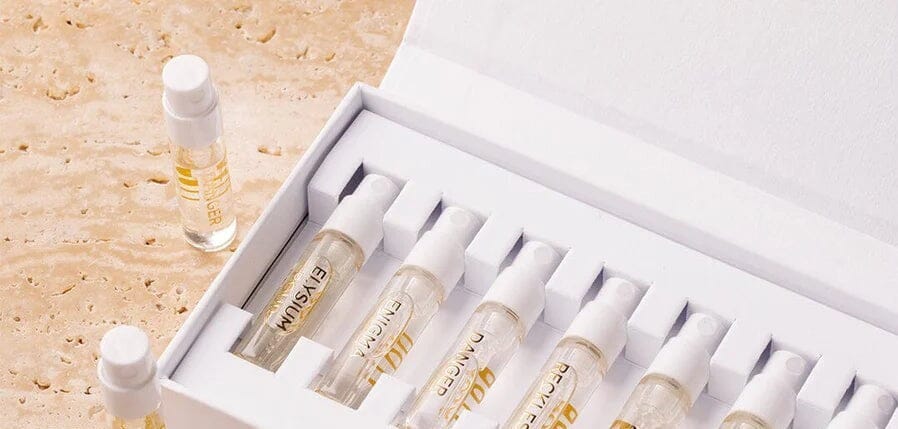 A white box from the Roja Parfums The Signatures Discovery Collection holds seven clear perfume sample vials with white caps and gold labels, embodying the spirit of olfactive art. One small vial from this elegant parfum set is placed outside the box on a beige textured surface.
