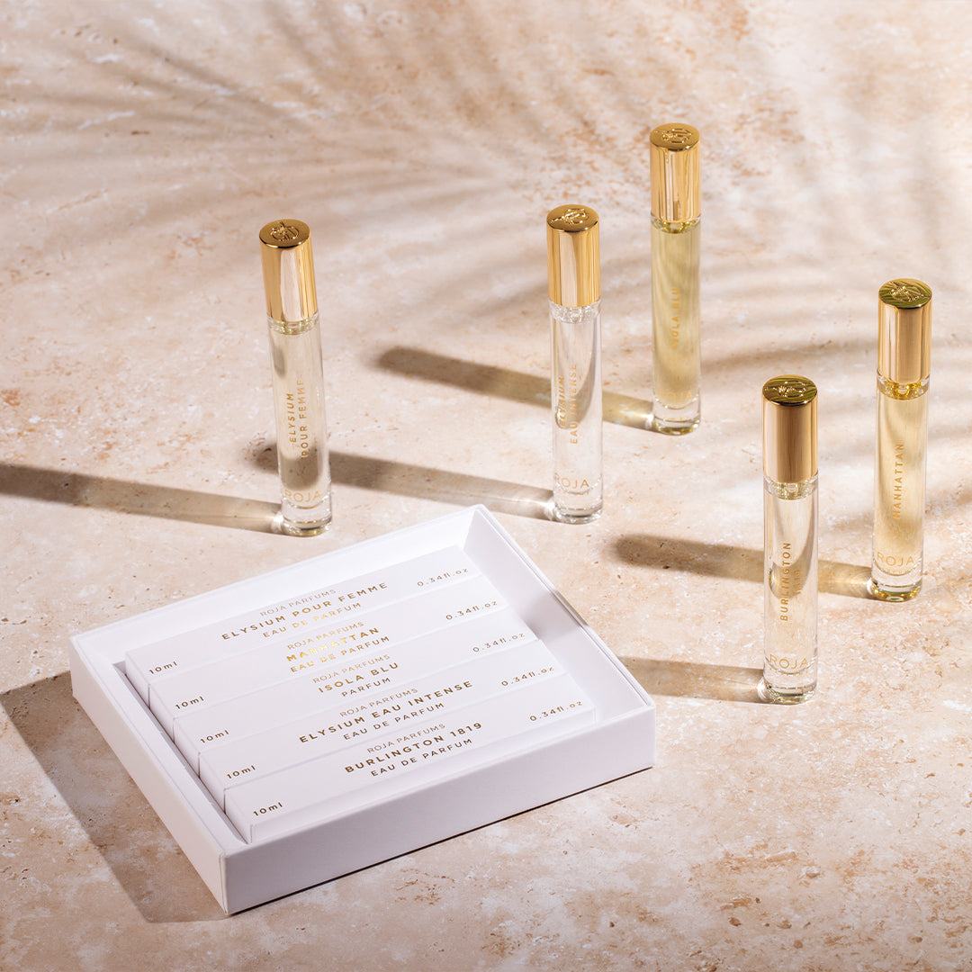 Five glass perfume vials with gold caps stand on a beige surface next to an open white box holding five more labeled perfume vials. Soft shadows and sunlight create a warm, elegant atmosphere.