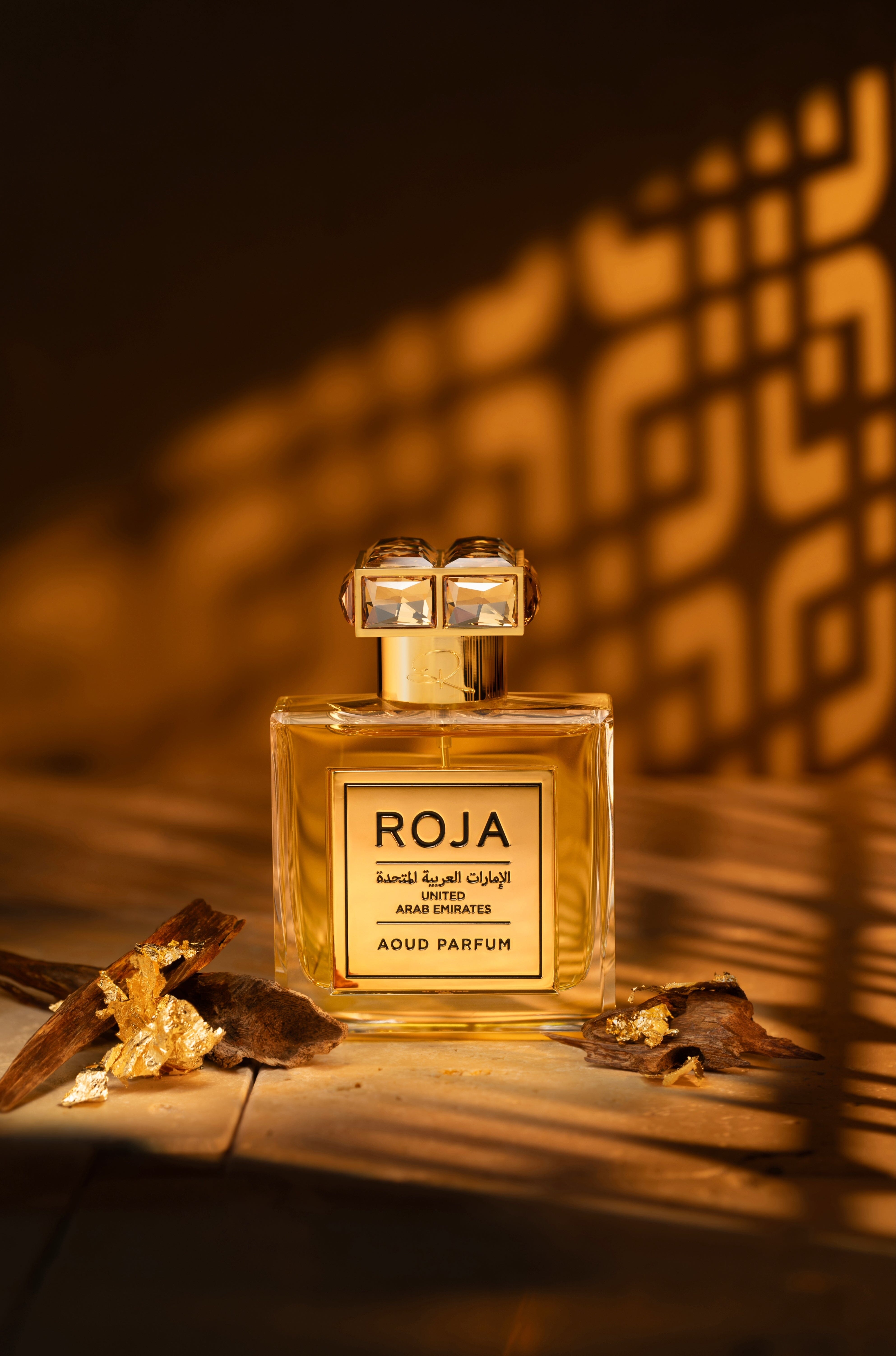 A bottle of Roja Parfums United Arab Emirates, a luxurious fragrance, stands on a wooden surface adorned with gold flakes and oud wood. Warm light illuminates the scene, casting geometric shadows on the wall behind it, while subtle hints of frankincense evoke an air of opulence and mystery.