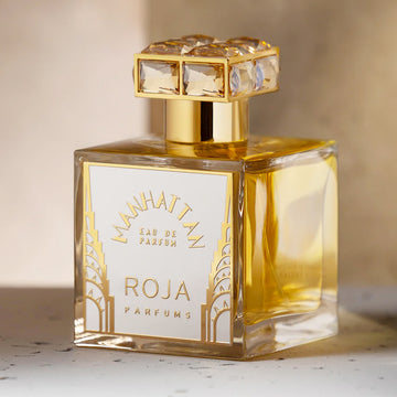 A clear glass bottle of Manhattan Eau de Parfum by Roja Parfums with gold accents and a jeweled square cap, filled with golden liquid, sits on a light surface.