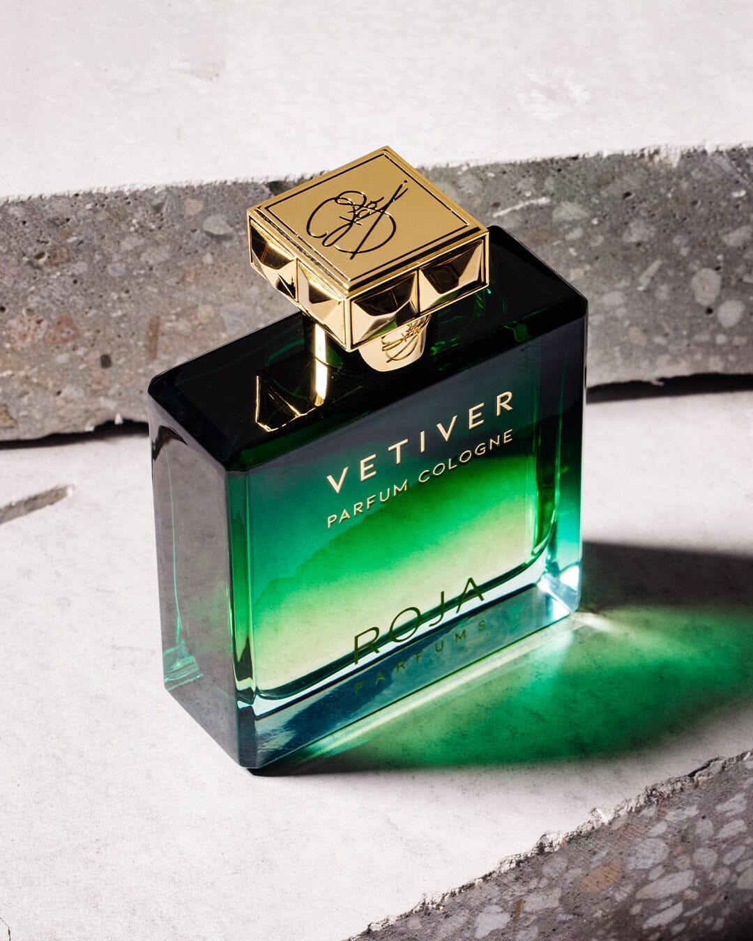 A green glass bottle of Vetiver Pour Homme by Roja Parfums, featuring a gold cap, rests on textured stone and casts a vibrant green reflection, epitomizing the brand's renowned masculine perfumery.