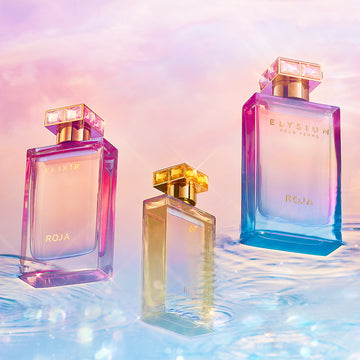 Three vibrant perfume bottles with gold caps, blending pink, purple, blue, and yellow hues, are displayed on a reflective watery surface against a pastel, sunset-colored sky background. The bottles are labeled ROJA.
